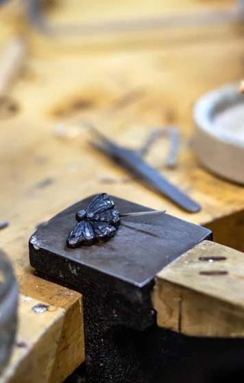 A close-up of a dark metal butterfly-shaped jewellery piece on a metal block, with an artist’s hand holding a fine brush to apply detail work. A close-up of a dark metal butterfly-shaped jewellery piece on a metal block, with an artist’s hand holding a fine brush to apply detail work.
